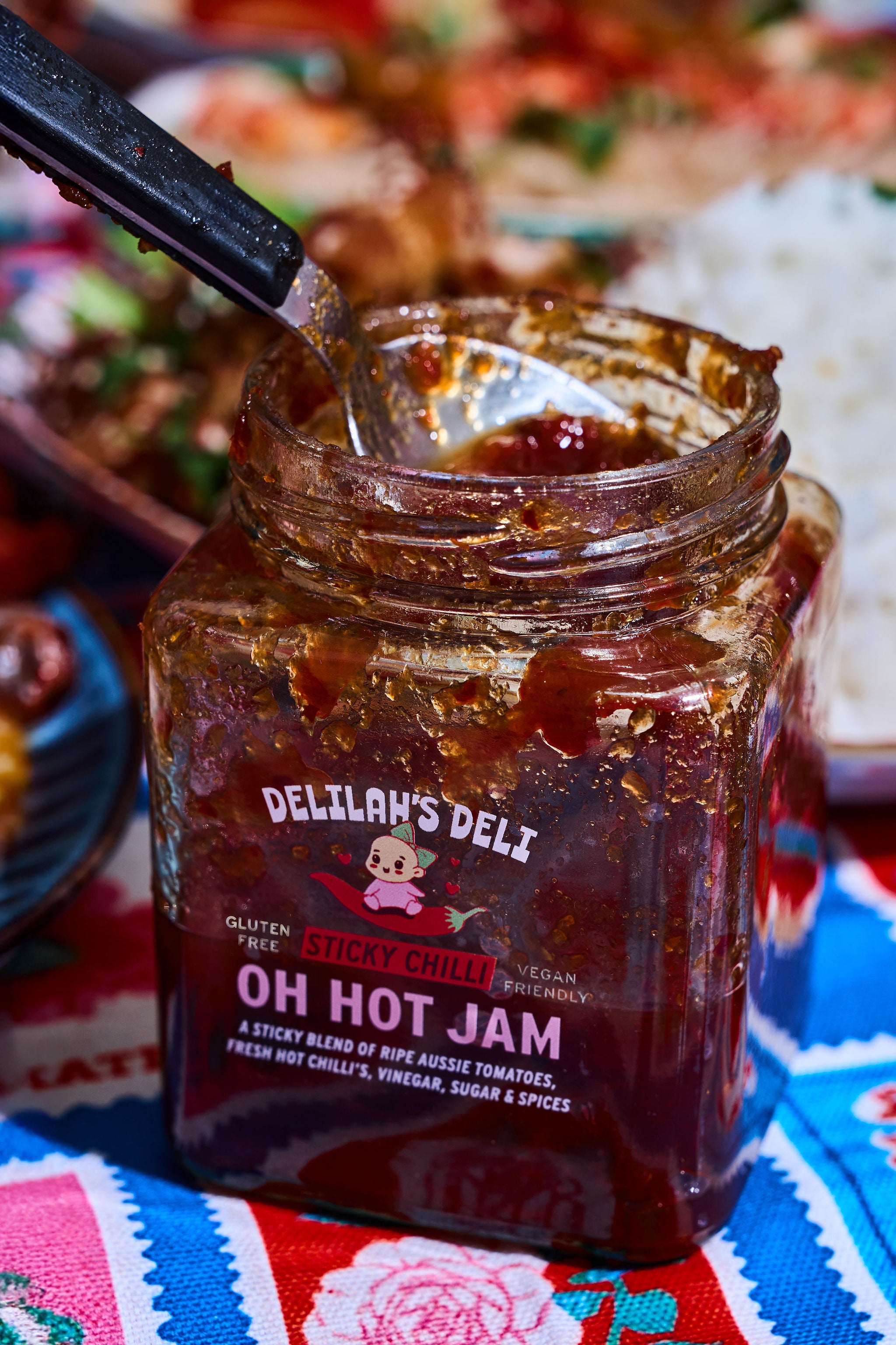 Jar of Delilah's Deli Oh Hot Jam on a colorful tablecloth with food in the background