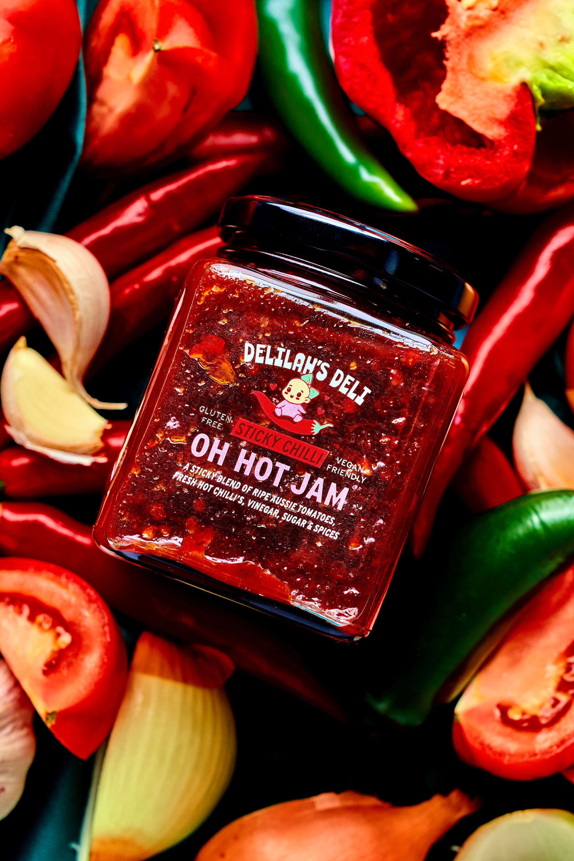 Jar of Delilah's Deli Oh Hot Jam surrounded by peppers, tomatoes, and garlic on a dark background