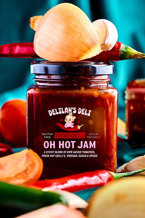 Jar of Delilah's Deli Oh Hot Jam surrounded by vegetables on a blue background