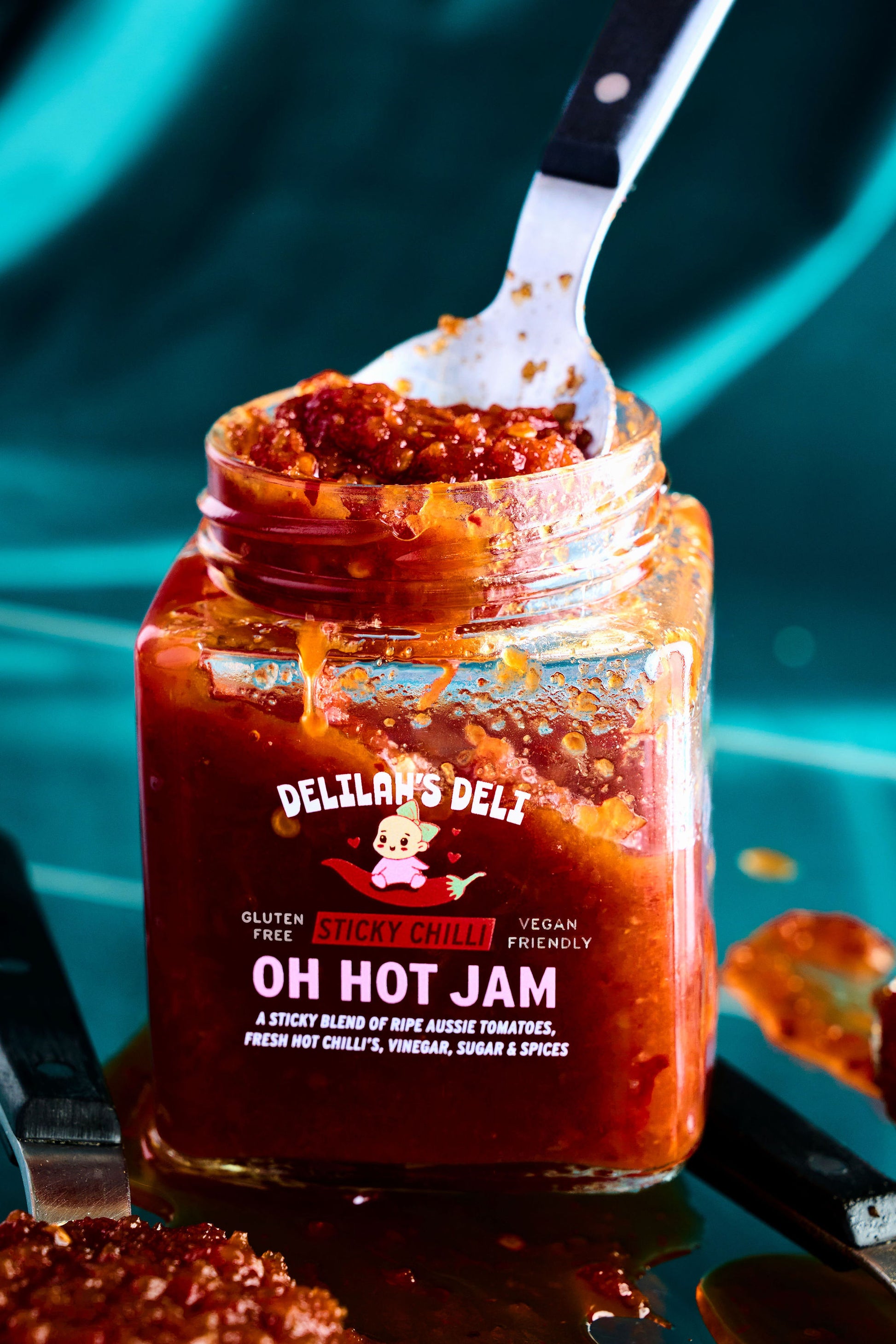 Jar of Delilah's Deli Oh Hot Jam with a spoon inside, on a teal background