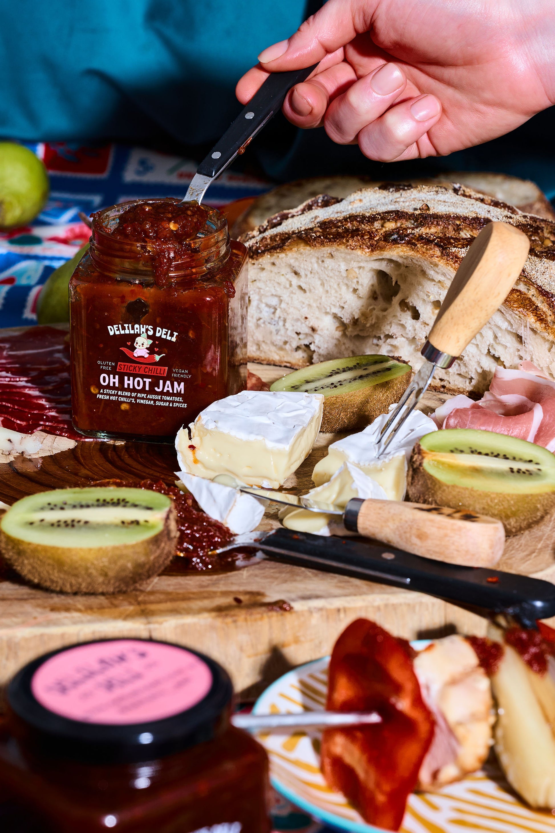 Person spreading jam on bread with a jar of 'Oh Hot Jam' and sliced kiwis on a wooden board.