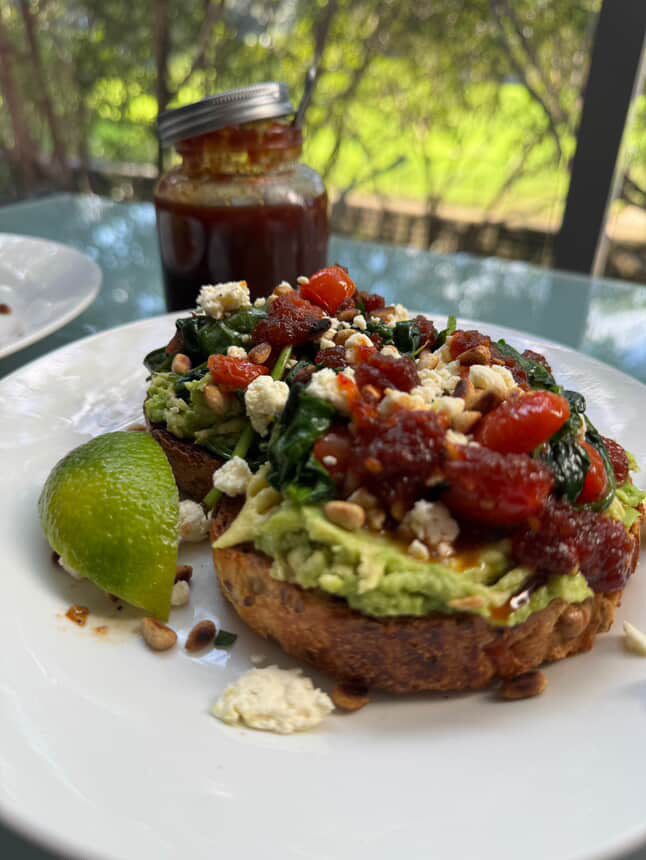 Avocado toast with toppings on a white plate with a jar of hot chilli jam in the background.