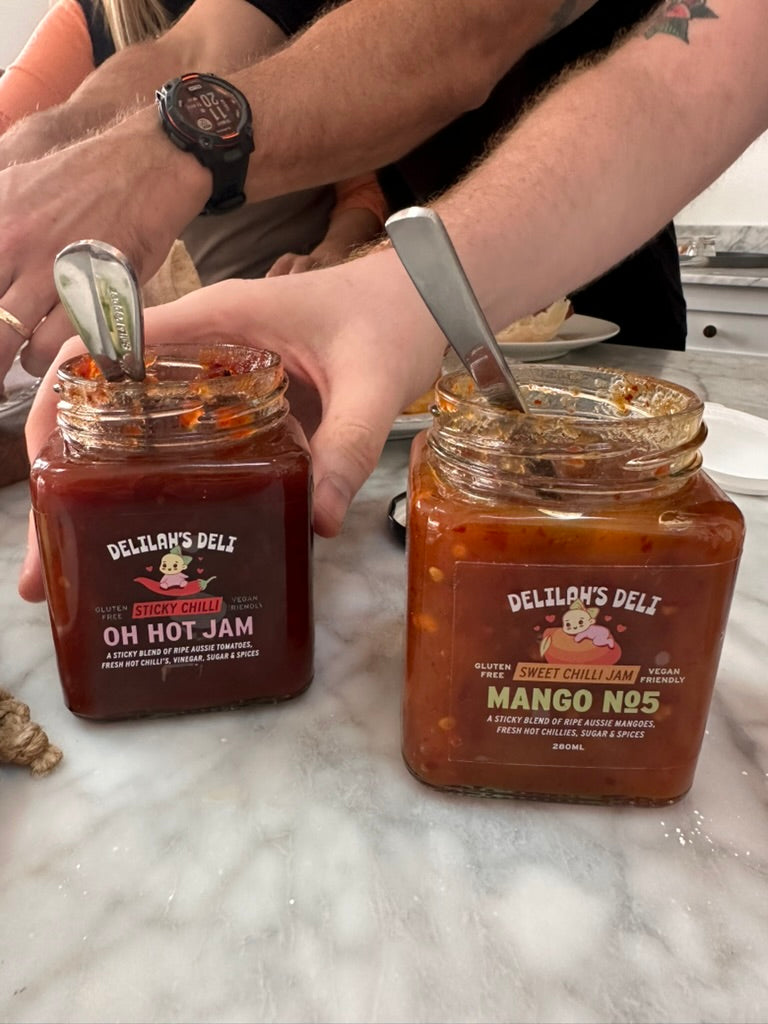 Two jars of Delilah's Deli jam with spoons on a marble surface.
