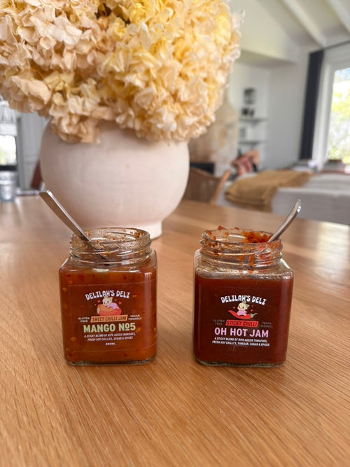 Two jars of Delilah's Deli jam on a table with a decorative flower arrangement in the background.