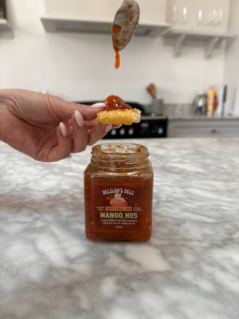 Hand holding a cracker with mango jam above a jar of Delilah's Deli mango jam on a kitchen counter.