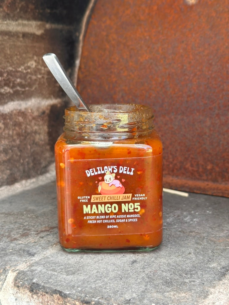 Jar of Delilah's Deli Mango N25 sweet chilli jam with a spoon on a rustic surface.