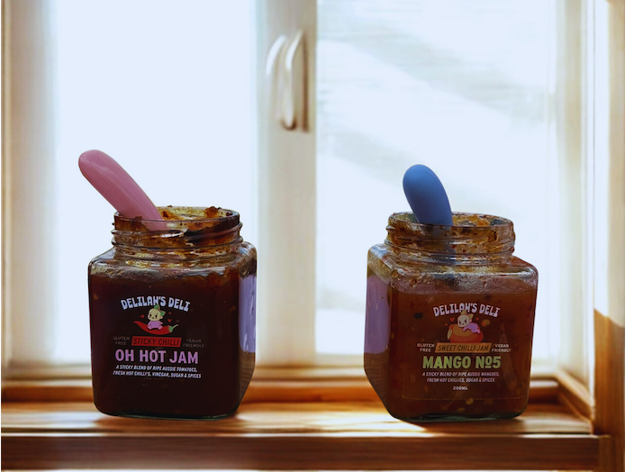 Two jars of Delilah's Deli jam on a windowsill with a pink and blue spoon.